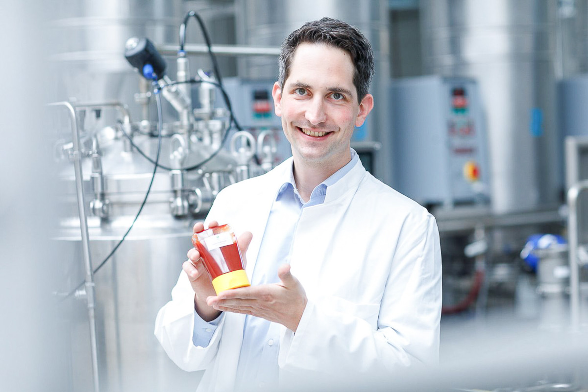 Mitarbeiter im Laborkittel mit einem Produkt vor Produktionsanlagen